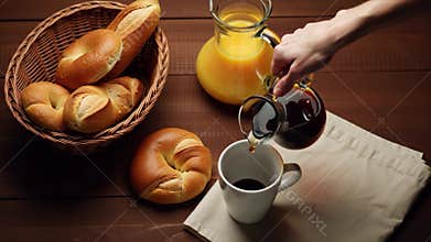 Pouring black coffee into white ceramic cup with various bread in woven basket and orange juice in transparent glass jar