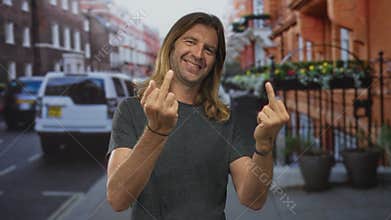 Blond man with long hair wearing dark tee shows double middle finger gesture on empty urban street outdoors; defiance