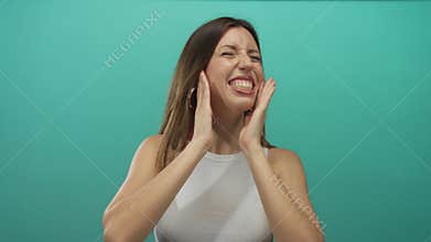 Young hispanic woman in white tank top grimacing while covering her ears in a teal studio for earache; discomfort