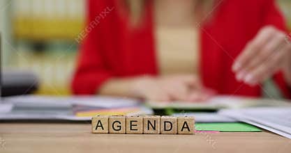 Wooden blocks spell Agenda word while woman turns pages at table
