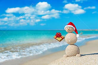 Smiley sandy snowman at beach in christmas hat with golden gift.