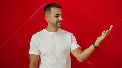 Young hispanic man pointing at his palm over an isolated red background looking amused and engaged