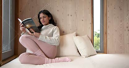 Woman relaxing at home, reading book by window on cozy day, copy space
