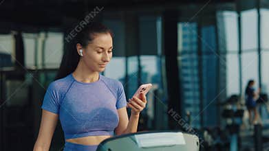 Woman in sportswear walks on treadmill checking smartphone