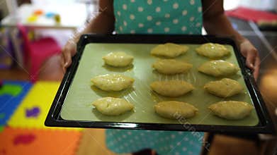 Housewife in an apron carries a tray of raw buns smeared with a beaten egg