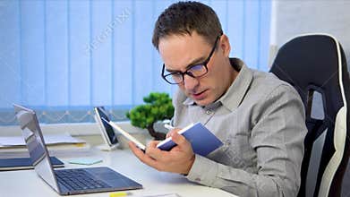 Image concentrated businessman in blue shirt holding and looking blue notebook during work in office. Tired worker reads