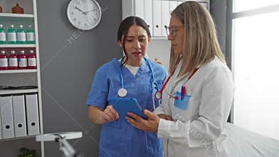 Women doctors in medical uniforms collaborate in a clinic using a tablet, examining patient information, in a professional indoor