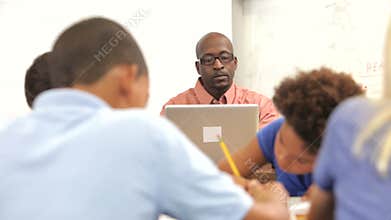 Teacher Using Laptop At Desk As Students Work