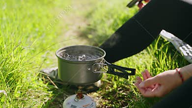 Woman on backpacking trip boils water in cooking pot on portable mini gas stove. Turns off burner outdoors. Ultralight