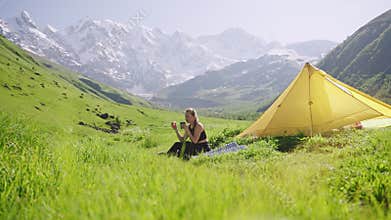 Woman enjoys breakfast, drinks coffee on outdoor backpacking trip with yellow tent. Scenic mountain landscape, green