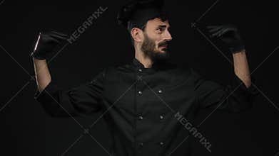 Hispanic male chef with a beard gestures expressively wearing black gloves and uniform against a dark background, conveying