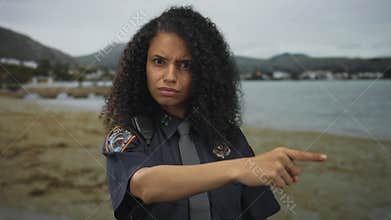 Woman police officer furrows brow and points finger on street by seaside town pier; authority command anger