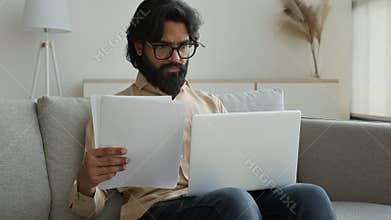 Indian muslim man freelancer working laptop at home Arabian businessman analyze documents paperwork read papers pay