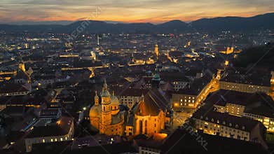 Aerial view of Graz, Austria at dusk, with illuminated streets and iconic Schlossberg hill. Fly over Cathedral of Saint