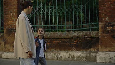 Mom in jeans and jacket and daughter walking on the street to the school. Side view of a happy child is holding parents