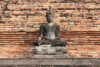 A stone statue of Buddha was installed in front of a brick wall in a park in Sukhothai (Thailand)