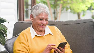 Happy senior woman enthusiastically uses smartphone on a comfortable couch indoors, smiling and laughing