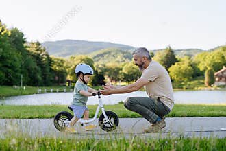 Father teaching toddler son to ride balance bike while wearing safety helmet.