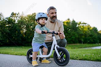Father teaching toddler son to ride balance bike while wearing safety helmet.