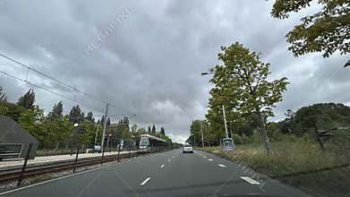 Cloudy urban road beside tram line with moving car, trees, and modern