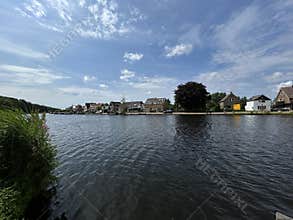 Wide riverside view with calm water, row of detached homes, trees, parked