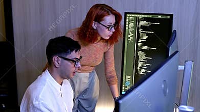 Two programmers collaborate at a dual-monitor workstation, reviewing and debugging code on screen in a modern office