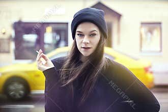 Young pretty woman smoking outside. Hipster outfit, wearing black hat and t-shirt, city yellow taxi on the background.