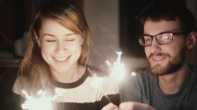 Slow motion. Young happy couple sitting at home in the evening and holding the sparklers, kissing each others.