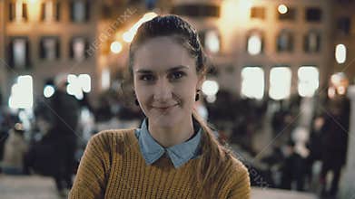 Portrait of young beautiful woman standing in the city centre in evening. Student girl looks at camera, smiling.