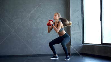 Young fitness woman doing squat with dumbbells in hands. Scene in the gym