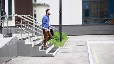 Young handsome bearded man in blue shirt and brown trousers is going outside. Student walking outdoors happily after