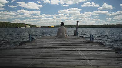 Brunette in plaid sad sitting on an old wooden pier and looks out at the water and forest in the distance.