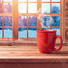 Red cup on wooden table over window with winter nature view. Winter and Christmas holiday concept. Cup mock up template for logo s