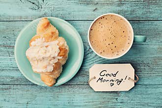 Coffee mug with croissant and notes good morning on turquoise rustic table from above, cozy and tasty breakfast