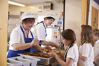 Two women serving food to a girl in a school cafeteria queue