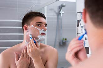 Handsome guy shaving his face with a razer and looking at the mi