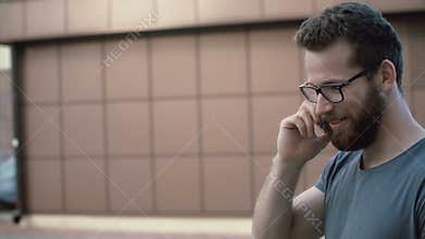 Portrait of young attractive man with beard and glasses walking on the street, using smarphone. Guy talking by the phone