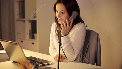 Woman with laptop calling on phone at night office