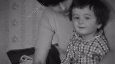 Mother dress shoes on little son in plaid shirt. Baby smile in camera. Woman play with child on bed.