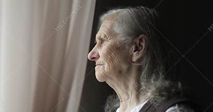 An old woman stands by the window and holds the curtain.