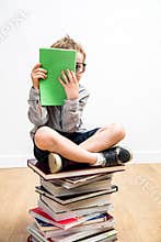Shy bookworm child sitting on many books hiding behind book