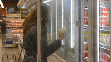 Young woman buying dairy or refrigerated groceries at the supermarket in the refrigerated section opening glass door of the fridge
