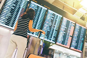 Asian woman traveler looking at flight information screen in an airport, holding suitcase, travel or time concept