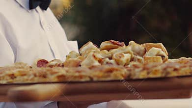 A young waiter in the restaurant serving banquet, delivers delicious Italian dishes on a large wooden plate, in slow