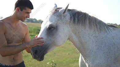 Young handsome man embracing and stroking white horse outdoors. Guy hugging a horse in the field, he caresses and