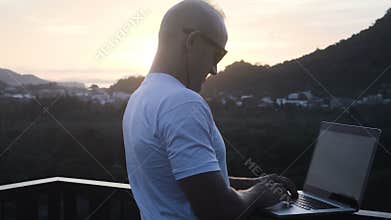 Outdoors portrait of handsome young man working on a laptop computer while standing on a rooftop over beautiful sunrise
