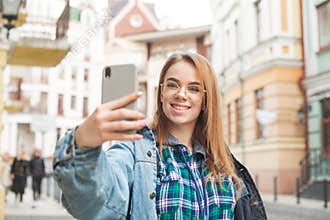Happy girl traveler in casual clothing makes a selfie on the streets town, smiles and looks at the camera of a smartphone. Girl