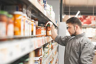 Man chooses canned vegetables on a shelf of a supermarket, holds a can of legumes and reads a label. Buyer selects canned