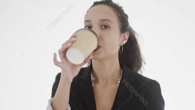 Appealing yound black-haired businesswoman with hoop earrings looks seductively straight at camera and makes a sip from