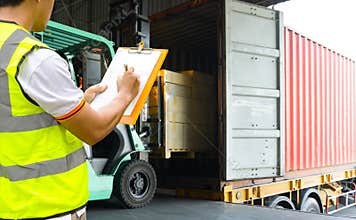 Warehouse worker holding clipboards control forklift loading shipment goods into container truck.